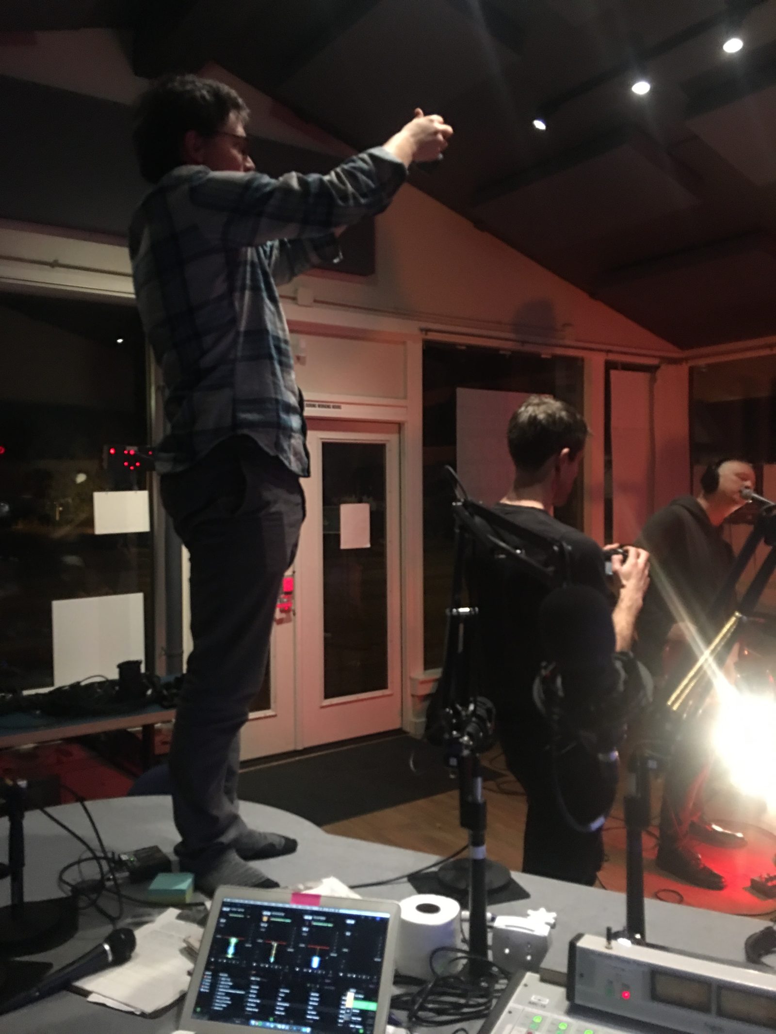 A radio studio at night: one person stands on the desk holding up a phone to film, another films beside the microphones, and a performer in headphones sings at a mic under red light.